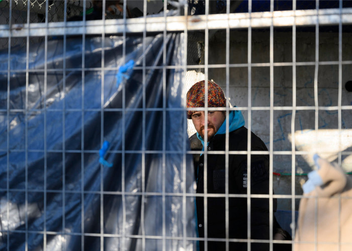A man, a refugee, is seen through the barrier of steel fencing.