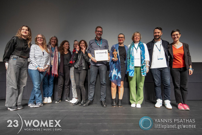 Group photo of various On the Move board members and representatives with a WOMEX award certificate and statuette.