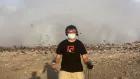 A young man in black t-shirt and medical face mask stands with two big audio recorders in his outstretched hands. Behind him is the brown wasteland of a rubbish tip, with birds rising up.