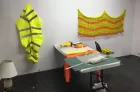 A simple studio space with strips of cut fabric draped over a desk. A yellow high-vis jacked hangs on the wall, next to a woven yellow and orange mat.
