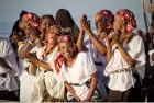 Half a dozen African women, outdoors, singing and smiling. They are dressed in identical white shirts and red and black headscarfs.