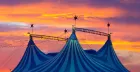 A three-peak circus tent, striped dark blue and teal, stands against a dusky orange sky.