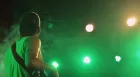 A musician wearing a guitar on a strap looks up at bright stage lights through a green haze.